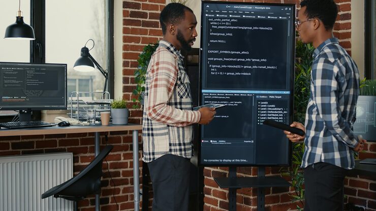 Photo team of software engineers leaving after analyzing source code on wall screen tv comparing errors using digital tablet. coders passing coworker programmer writing code for artificial intelligence.