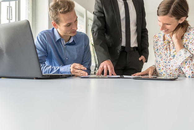 Photo team of three business people working together reviewing documents checking their data on laptop and digital tablet while at their white office desk