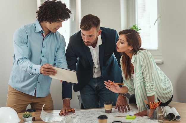 A team of three young architects is using digital tablet and communicating while analyzing blueprints of a new project in the office.