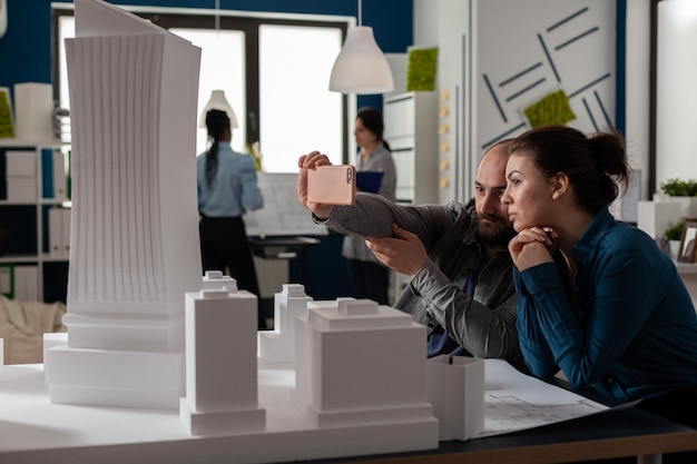 Team of two architect colleagues using smartphone in videocall conference with client presenting desk with buildings models. Architectural engineers working together with smart phone and maquette.