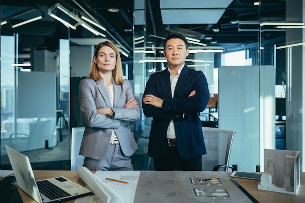 A team of two business people an Asian man and a woman look confidently at the camera serious and focused with their hands folded discussing the construction of a new business center