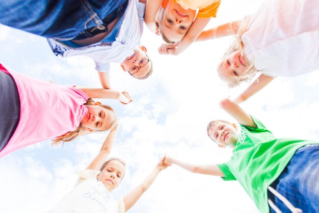 Team van lieve kinderen die nuttig tijd doorbrengen in een zomerkamp. Jonge schoolkinderen die in een cirkel staan, hand in hand op een hemelachtergrond