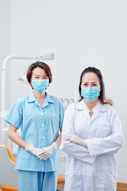 Team van positieve tandarts en jonge verpleegster in beschermende maskers die in de tandheelkundige kliniek staan