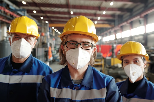 Team van productiemedewerkers met veiligheidshelmen en ademhalingsmaskers die in een grote werkplaats staan en naar de camera kijken