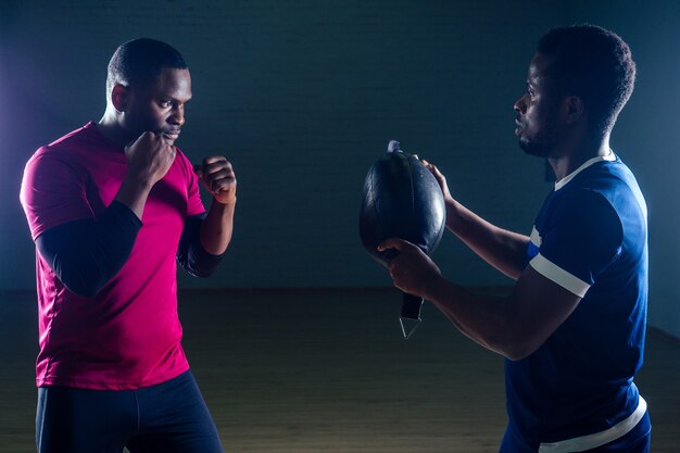 Lavoro di squadra due bei sportivi afroamericani nel pugilato su uno sfondo nero in palestra, allenatore e reparto che praticano la boxe