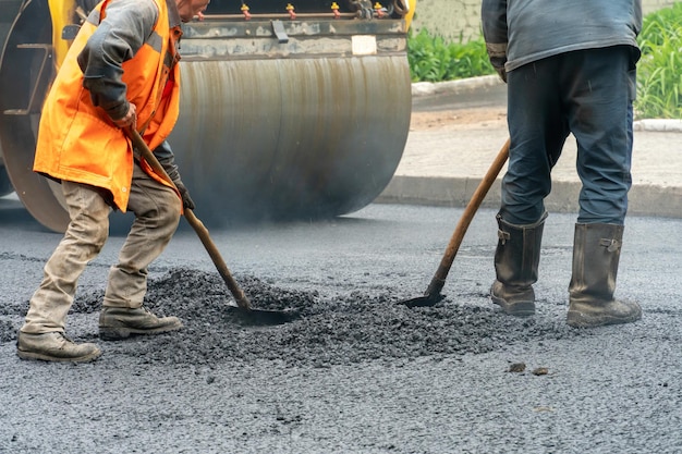 A team of workers with shovels is working on laying new asphalt\
road repairs workers with tools next to a roller asphalt stacker on\
the road during work