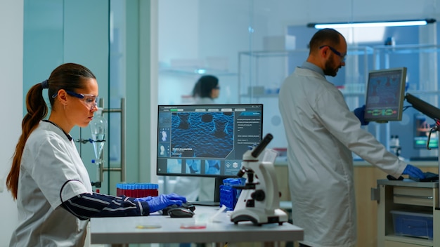 Team working to develop a new vaccine in modern equipped laboratory, woman typing at computer. Group of biochemistry scientists examining virus evolution using high tech for researching treatment