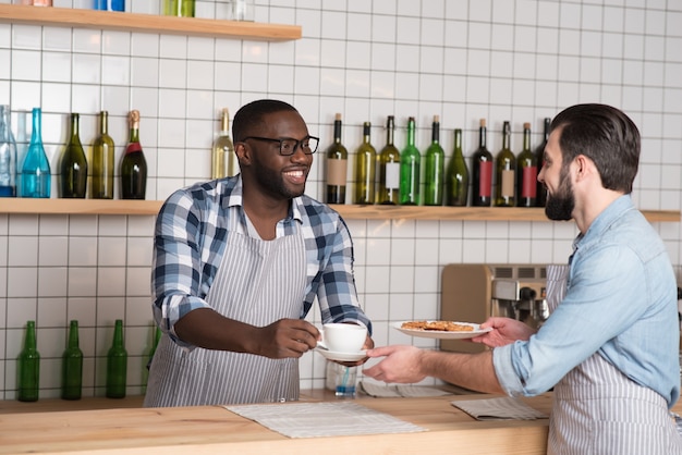 Teamwerk. Knappe vriendelijke behulpzame ober die aan de bar staat terwijl hij een kopje koffie geeft aan zijn vrolijke verantwoordelijke collega