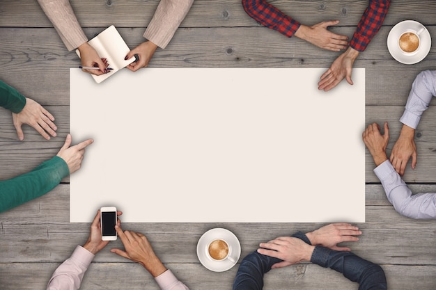 Photo teamwork and cooperation concept - top view of six people drawing or writing on a large white blank sheet of paper on wooden table.