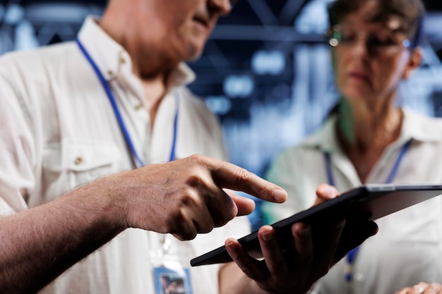 Teamworking colleagues using tablet to examine supercomputers for power fluctuations. Licensed engineers monitoring high tech server hub machines, identifying potential defects