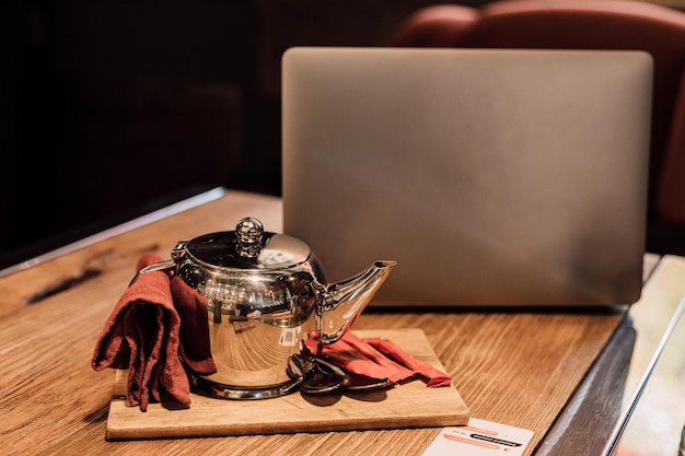 A teapot with tea stands near a laptop on a wooden table in a cafe