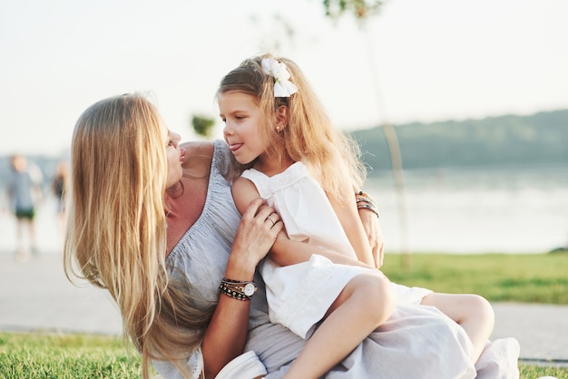 Teasing. Photo of young mother and her daughter having good time on the green grass with lake at background.