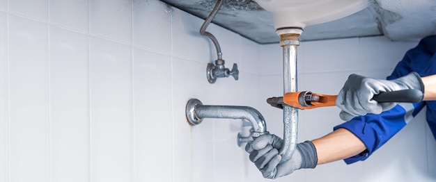 Technician plumber using a wrench to repair a water pipe under\
the sink concept of maintenance fix water plumbing leaks replace\
the kitchen sink drain cleaning clogged pipes is dirty or\
rusty