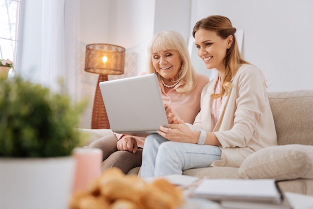 Dispositivo tecnologico. donne piacevoli positive allegre che si siedono insieme e che sorridono mentre usando un computer portatile