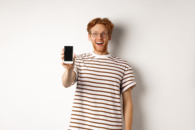 Technologie en e-commerce concept. Gelukkig jonge roodharige man in glazen met leeg smartphonescherm, camera kijken verbaasd, staande op een witte achtergrond.
