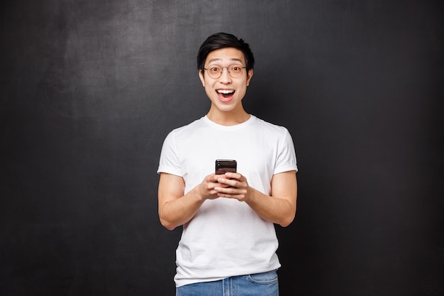 Technologie, gadgets en mensen concept. Super blij lachend positieve Aziatische man in bril en t-shirt, reageer geamuseerd en vroeg zich af over geweldig nieuws ontvangen via e-mail, houd mobiele telefoon camera