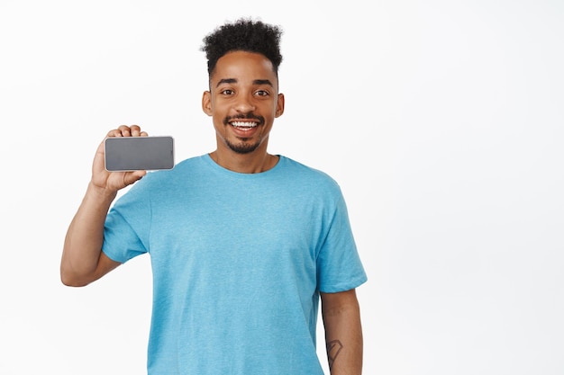 Technologie. Glimlachende Afro-Amerikaanse man met smartphone horizontaal scherm, mobiele telefoon interface, app aanbevelen, staande in blauw t-shirt tegen witte achtergrond.