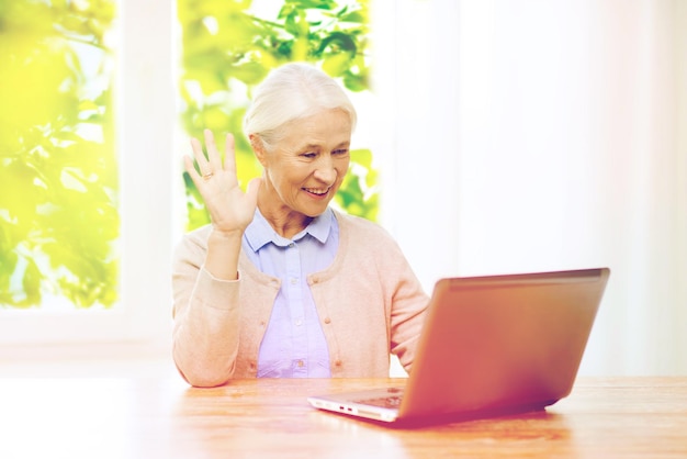 technologie, leeftijd en mensenconcept - gelukkige senior vrouw met laptopcomputer die thuis videochat heeft en met de hand over het raam zwaait met groene natuurlijke achtergrond