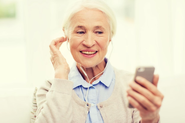 technologie, leeftijd en mensenconcept - gelukkige senior vrouw met smartphone en oortelefoons die thuis naar muziek luisteren