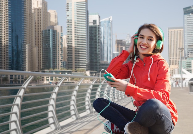 technologie, levensstijl en mensenconcept - lachende jonge vrouw of tienermeisje met smartphone en koptelefoon luisteren naar muziek over de straatachtergrond van de stad Dubai