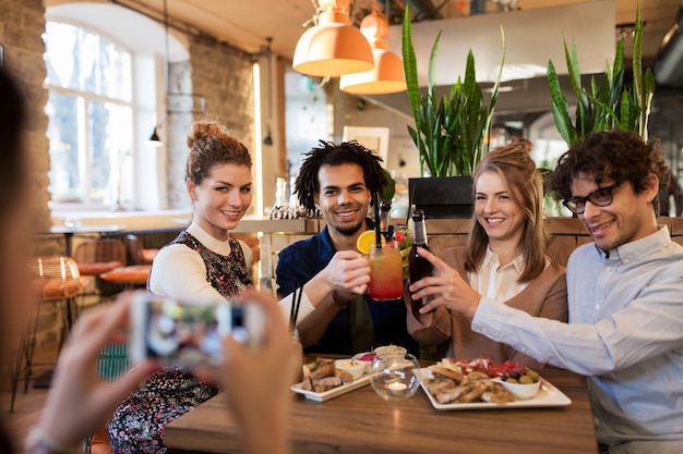 technologie, levensstijl, vakantie en mensenconcept - gelukkige vrienden die drankjes rammelen en fotograferen via smartphone in bar of café