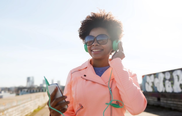 technologie, lifestyle en mensen concept - lachende Afro-Amerikaanse jonge vrouw of tienermeisje met smartphone en koptelefoon luisteren naar muziek buitenshuis