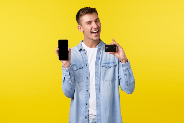 Technologie, lifestyle en reclame concept. Knappe lachende man introduceert nieuwe functie in app, met creditcard en mobiele telefoonscherm met blij gezicht, gele achtergrond.