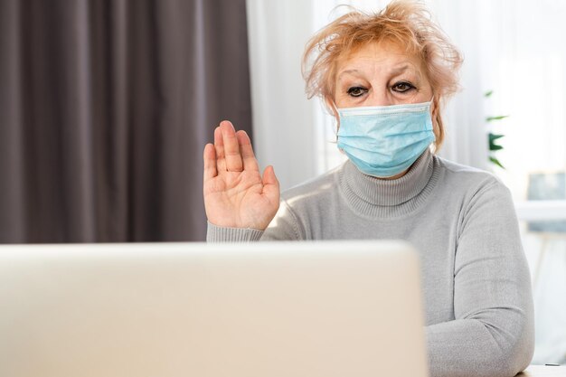 Technologie, ouderdom en mensenconcept - eenzame, trieste oudere oudere vrouw met een medisch gezichtsmasker dat thuis werkt en een videogesprek voert met een laptopcomputer tijdens de pandemie van het coronavirus COVID19. Blijf thuis