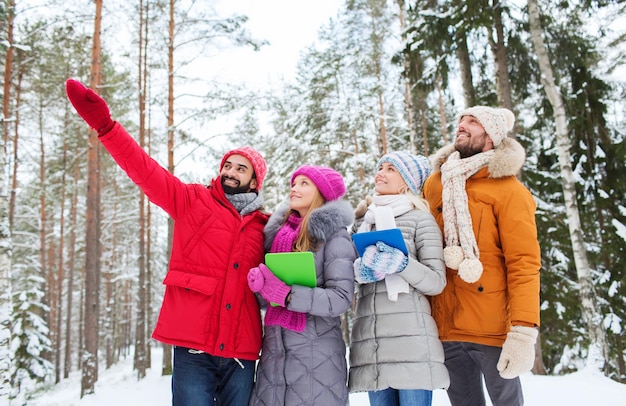 technologie, seizoen, vriendschap en mensenconcept - groep glimlachende mannen en vrouwen met tablet-pc-computers die met de vinger wijzen in het winterbos