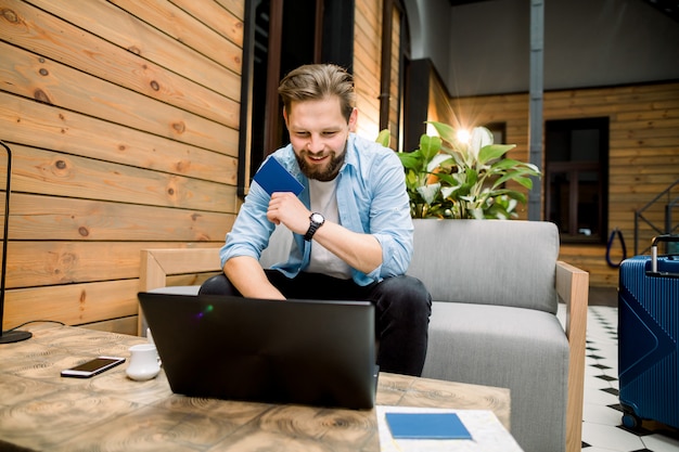 Technologie, winkelen, bankieren, reizen en lifestyle concept - knappe jonge blanke man met laptopcomputer en paspoort zitten in een moderne hotelhal of naaiatelier