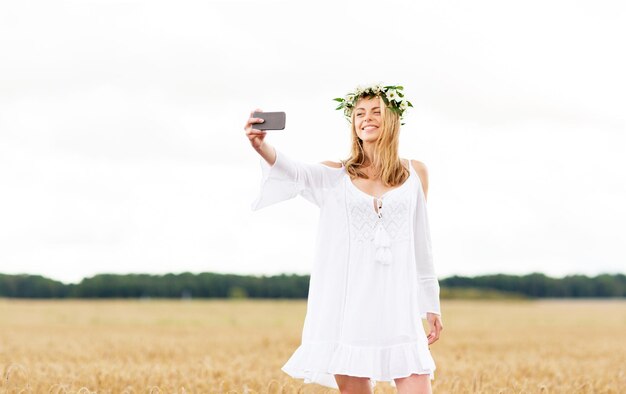 technologie, zomervakantie, vakantie en mensenconcept - lachende jonge vrouw in bloemenkrans die selfie maakt met smartphone op graanveld