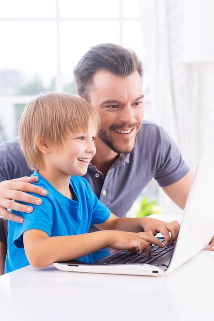 Technologies become easier. Cheerful father and son looking at the laptop while little boy typing something on keyboard and smiling