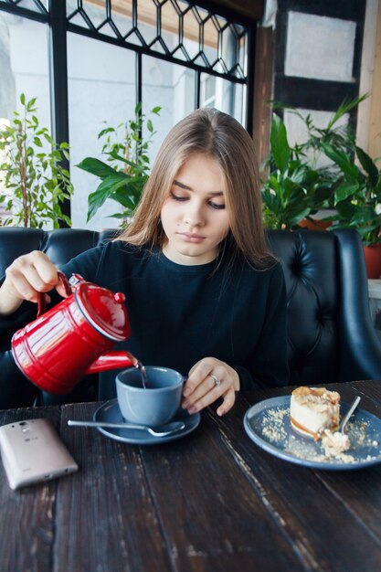 テクノロジー、感情、ライフスタイル、人々、十代の若者たちのコンセプト-モダンなコーヒーショップのインテリアに座っている間、彼女の携帯電話で読んでいる若い幸せな女性、美しい笑顔の女の子。お茶を飲む