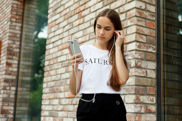 Technologies, emotions, people, music, beauty, fashion and lifestyle concept - Young woman with headphones dangling on her mobile phone as she walks in an urban street, view from a height