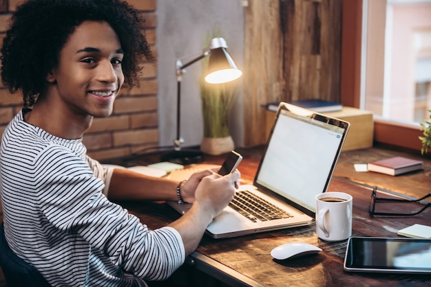 Technologies making life easier. Side view of cheerful young African man holding mobile phone and looking at camera with smile while sitting at his working place