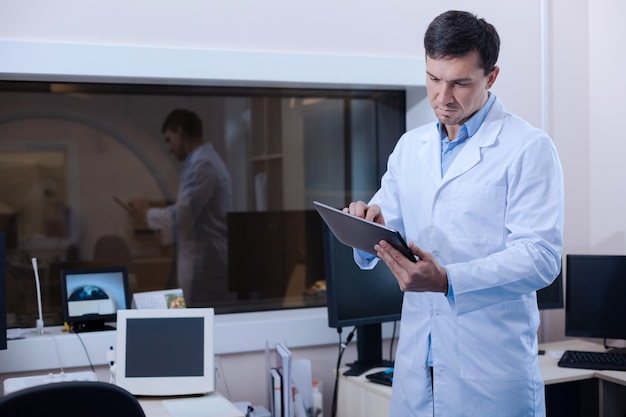 Technologisch apparaat. Professioneel ervaren goed uitziende arts die een tablet vasthoudt en deze gebruikt terwijl hij in het laboratorium staat