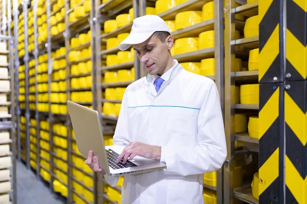 Technologist in a white coat with a laptop in his hands is in the warehouse of cheese in the shop for the production of butter and cheese quality control at the dairy plant racks with cheese