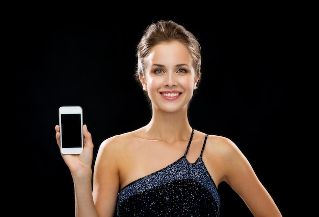 Photo technology, advertisement and lifestyle concept - smiling woman in evening dress with blank smartphone screen over black background