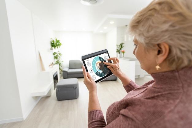 technology, automation and efficiency concept - smiling senior woman in glasses with tablet computer using smart home app over grey background.