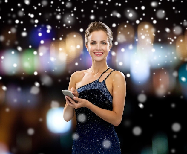 Tecnologia, natale, vacanze e concetto di persone - donna sorridente in abito da sera che tiene smartphone su luci notturne e sfondo di neve