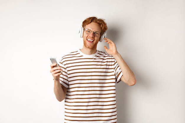 技術コンセプト。赤い髪とひげを持つ若い男がヘッドフォンで音楽を聴き、スマートフォンを使用して、カメラに微笑んで、白い背景。