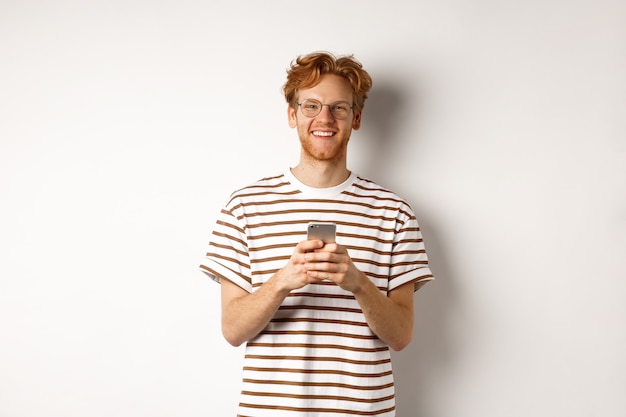 Концепция технологии и электронной коммерции. Рыжий парень в очках с помощью мобильного телефона и улыбается. Молодой человек со смартфоном, глядя счастливым в камеру, на белом фоне