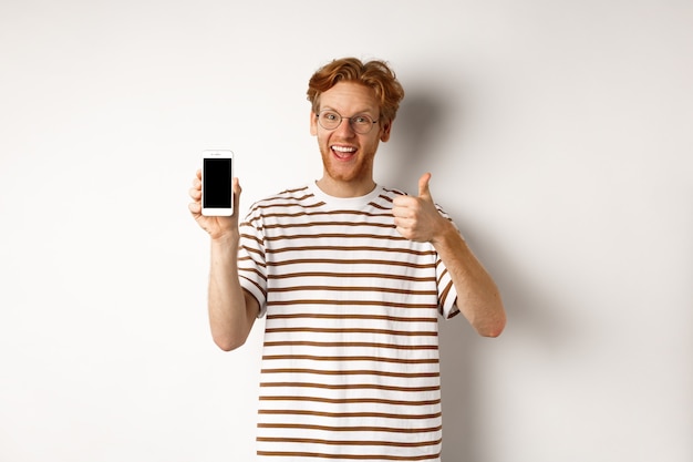 Technology and e-commerce concept. young man with red hair showing thumbs-up and blank smartphone screen, recommending app, white background.