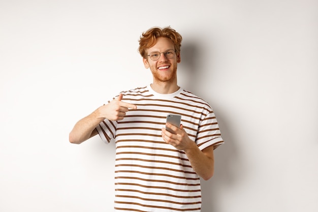 Foto tecnologia e concetto di e-commerce. giovane con i capelli rossi disordinati, con gli occhiali e la maglietta, puntando il dito sullo smartphone e sorridendo soddisfatto, sfondo bianco