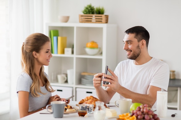 technology eating and people concept happy couple with smartphones having breakfast at home