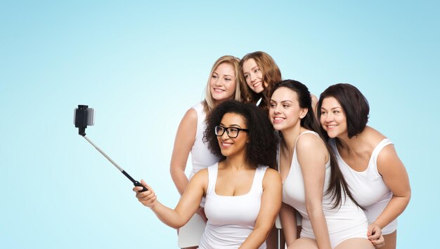 Foto tecnologia, amicizia, corpo positivo e concetto di persone - gruppo di donne felici in biancheria intima bianca che scattano foto con smartphone su selfie stick su sfondo blu