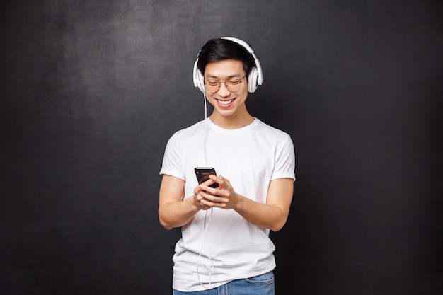 Concetto di tecnologia, gadget e persone. giovane uomo asiatico sorridente felice bello in maglietta, ascolta musica in cuffia, seleziona playlist nel telefono cellulare, amico sms,