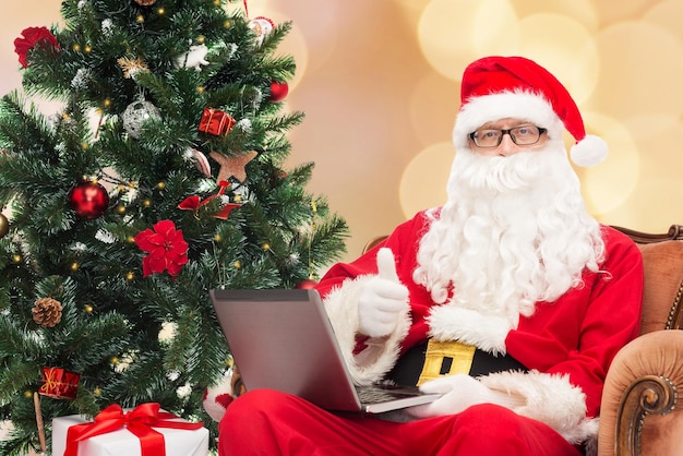 technology, holidays and people concept - man in costume of santa claus with laptop computer, gifts and christmas tree sitting in armchair over beige lights background