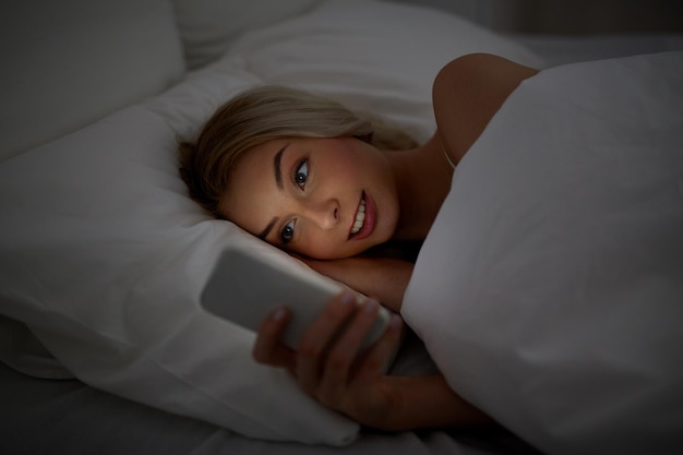 технологии, интернет, общение и концепция людей - счастливая улыбающаяся молодая женщина пишет смс на смартфоне в постели в домашней спальне ночью