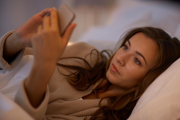 технологии, интернет, общение и концепция людей - счастливая улыбающаяся молодая женщина пишет смс на смартфоне в постели в домашней спальне ночью